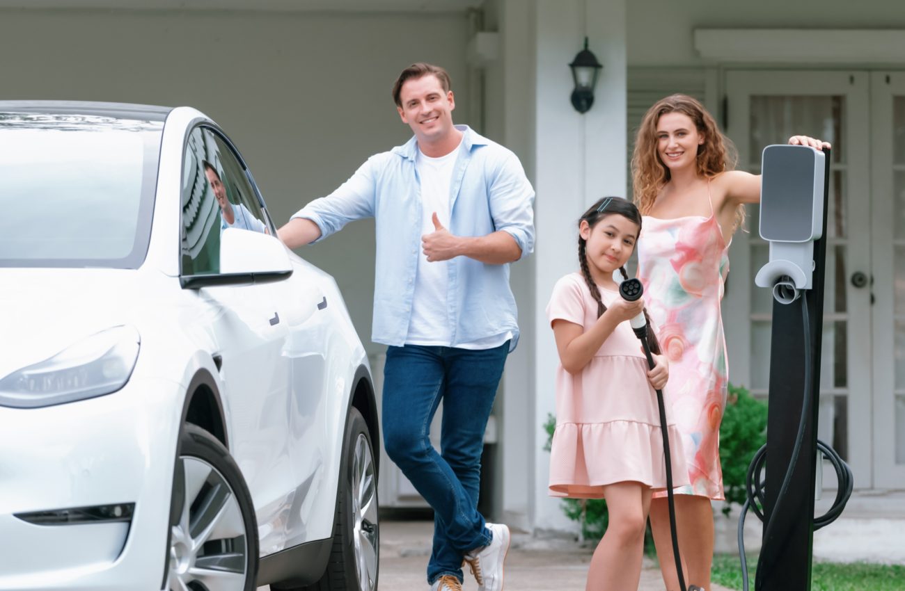 Modern family with environmental conscious recharging EV car with home EV charging station, Happy family with little young girl holding and pointing EV charger at camera. Synchronos